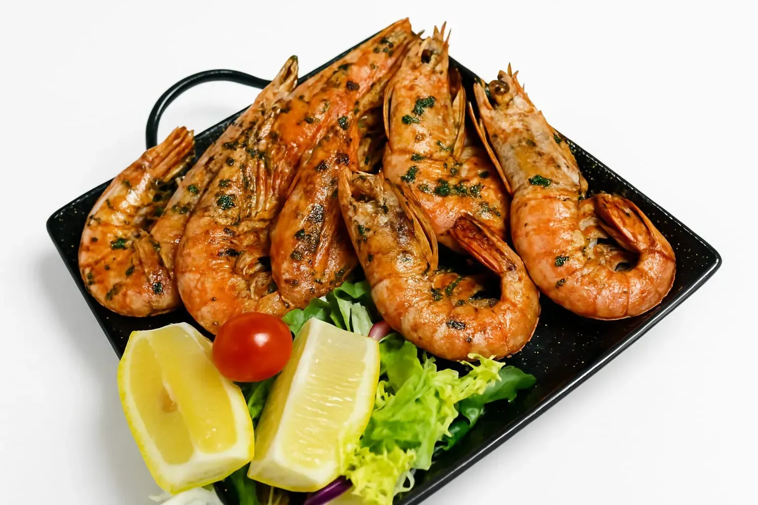 Gambas a la parrilla sazonadas con hierbas, servidas en un plato negro con rodajas de limón, un tomate cherry y lechuga fresca.