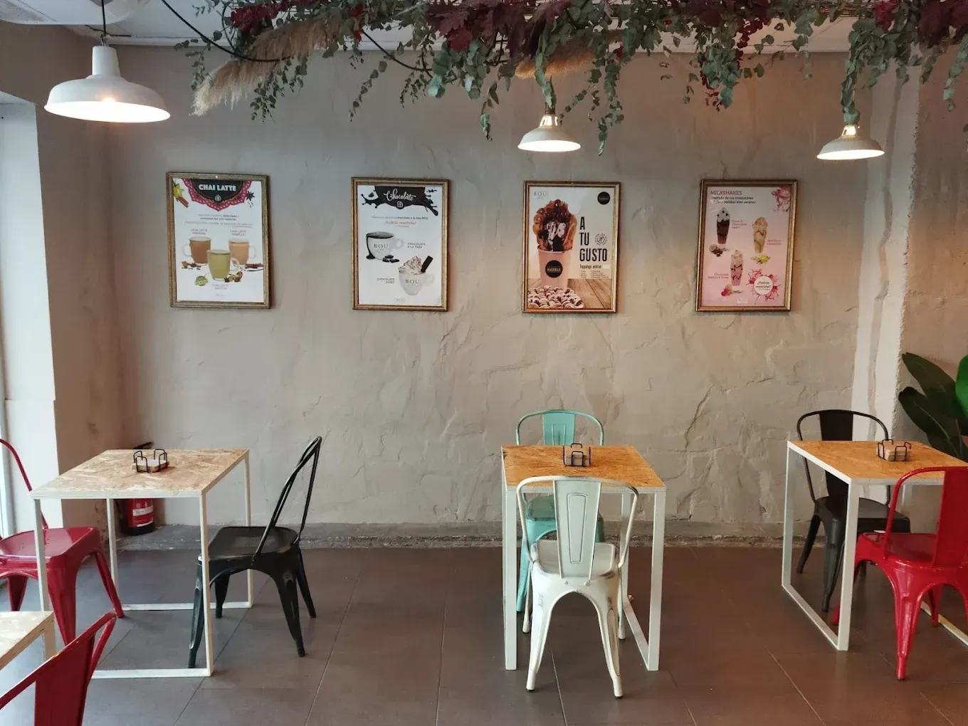 El interior de una cafetería cuenta con tres mesas pequeñas, sillas de metal de colores y carteles con el menú enmarcados sobre una pared texturizada de color beige.