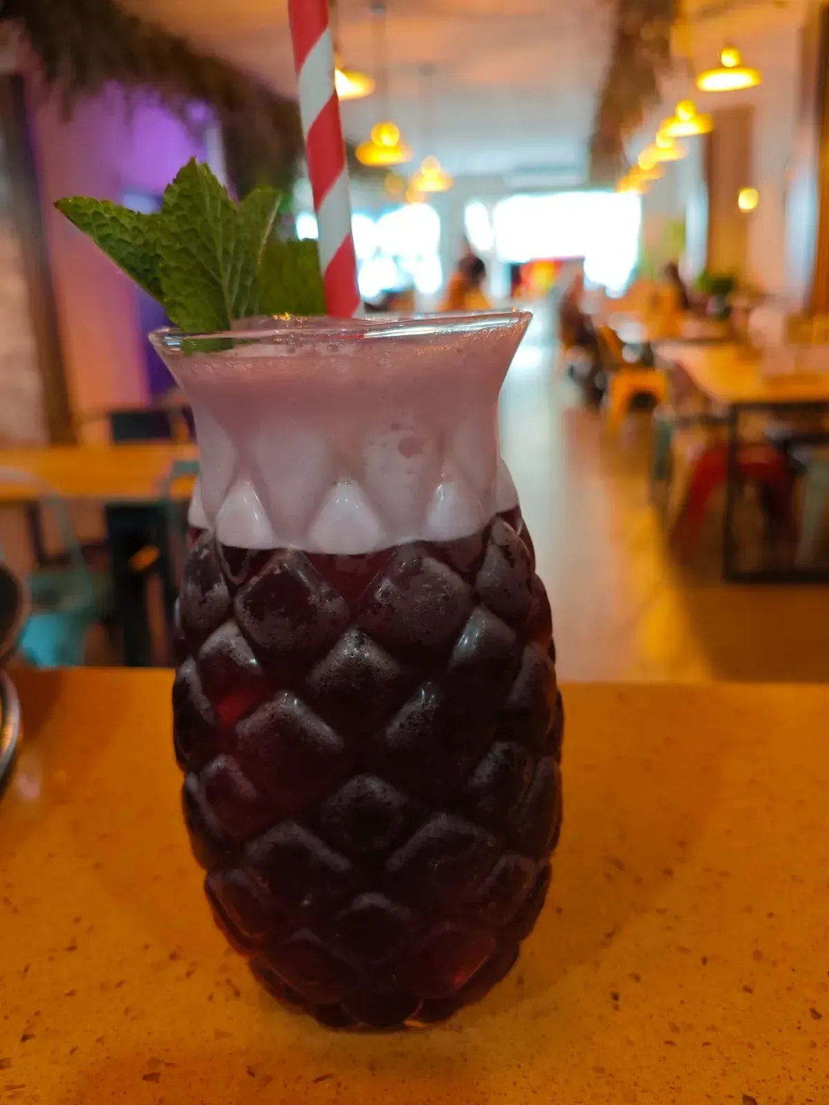 Un cóctel oscuro en un vaso con forma de piña, coronado con menta y una pajita roja y blanca, sobre una mesa en un restaurante borroso.