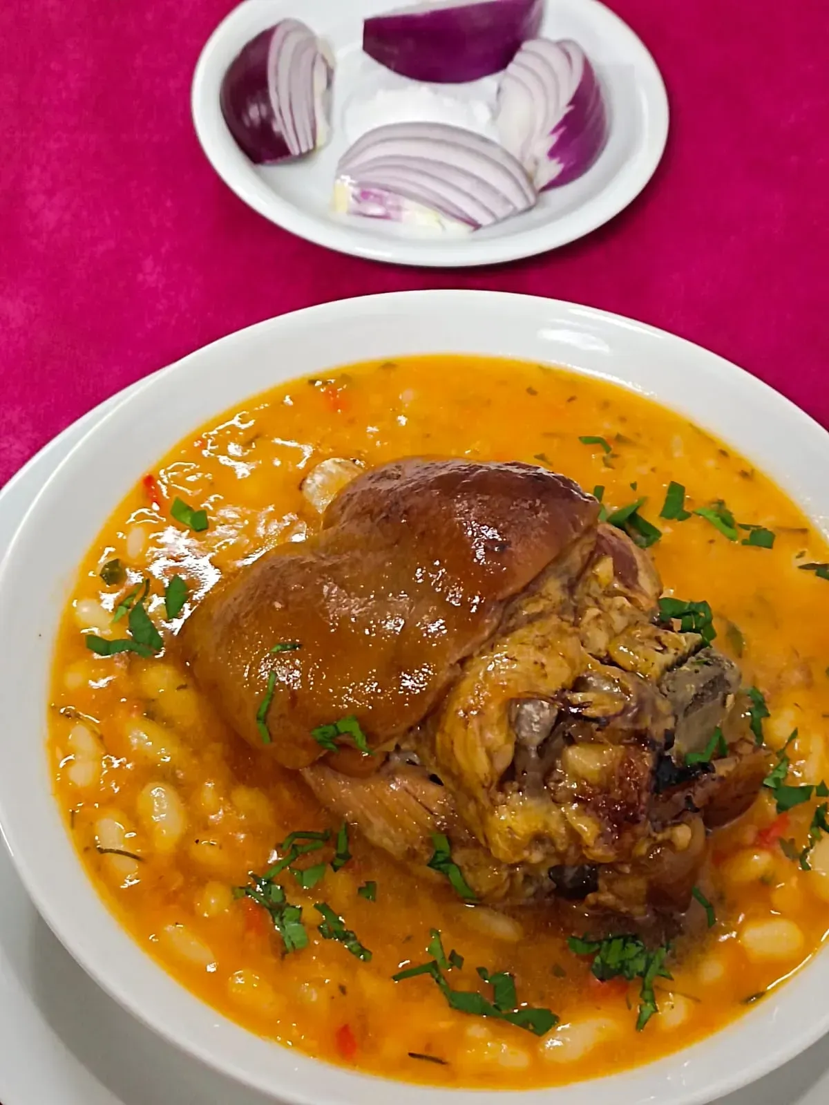 Un tazón de estofado de alubias blancas cubierto con un codillo de cerdo estofado, servido con una guarnición de rodajas de cebolla roja sobre una superficie rosada.
