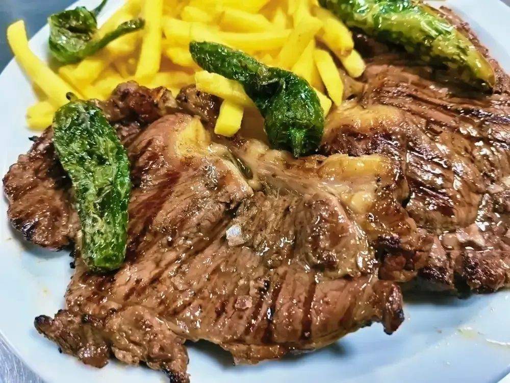 Un filete a la parrilla servido en un plato blanco con patatas fritas doradas y varios pimientos shishito verdes asados.