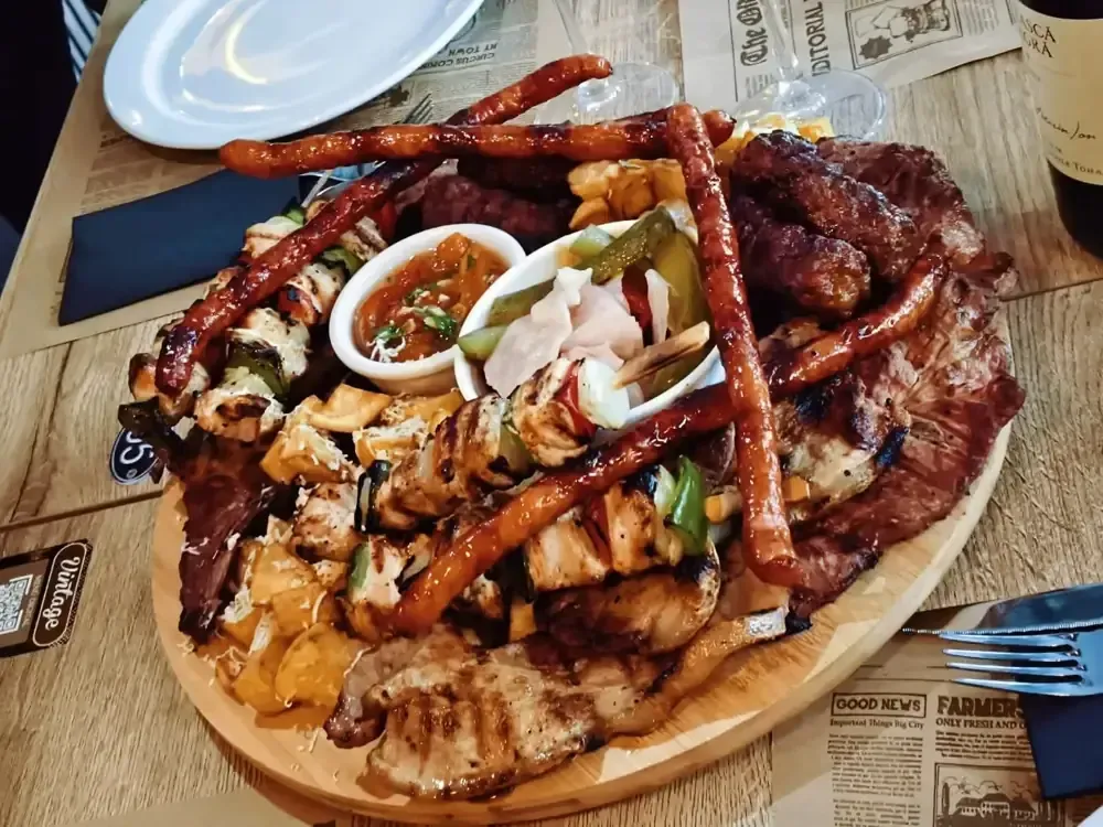 Una tabla de madera para servir repleta de diversas carnes a la parrilla, brochetas, salchichas, un pequeño cuenco de salsa y verduras encurtidas.