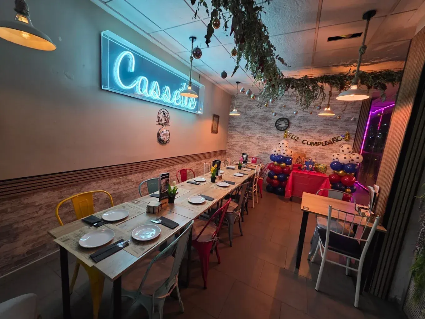 El interior de un restaurante con una mesa larga de madera, sillas coloridas y un letrero de neón azul con la palabra 