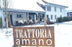 An einem schneebedeckten Tag steht vor einem weissen, zweistöckigen Restaurantgebäude ein rustikales Schild mit der Aufschrift „TRATTORIA amano“.
