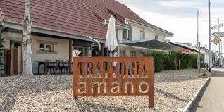 Vor einem weissen Gebäude mit Terrasse und braunem Ziegeldach steht ein verwittertes Metallschild mit der Aufschrift „TRATTORIA amano“.