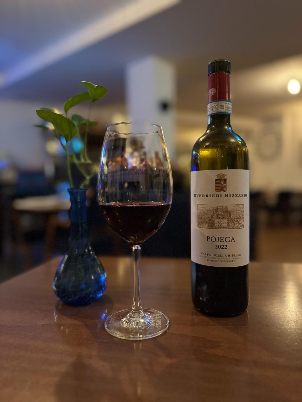 Auf einem Holztisch stehen ein Glas Rotwein, eine Flasche und eine blaue Vase mit Grünzeug.
