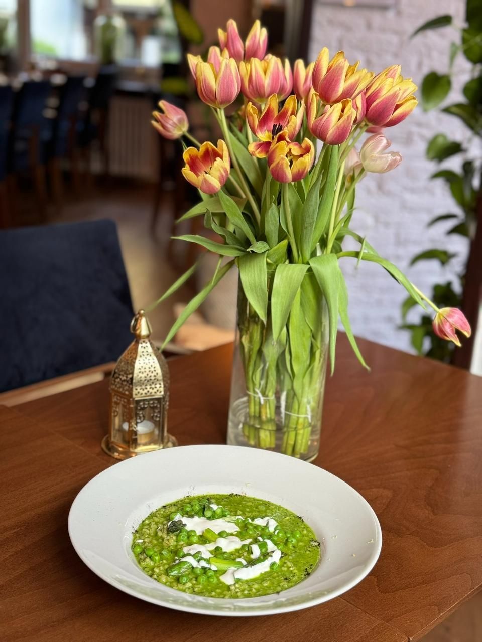 Ein grünes, pfannkuchenähnliches Gericht mit weissem Zuckerguss auf einem weissen Teller, Tulpen in einer Vase, auf einem Holztisch.