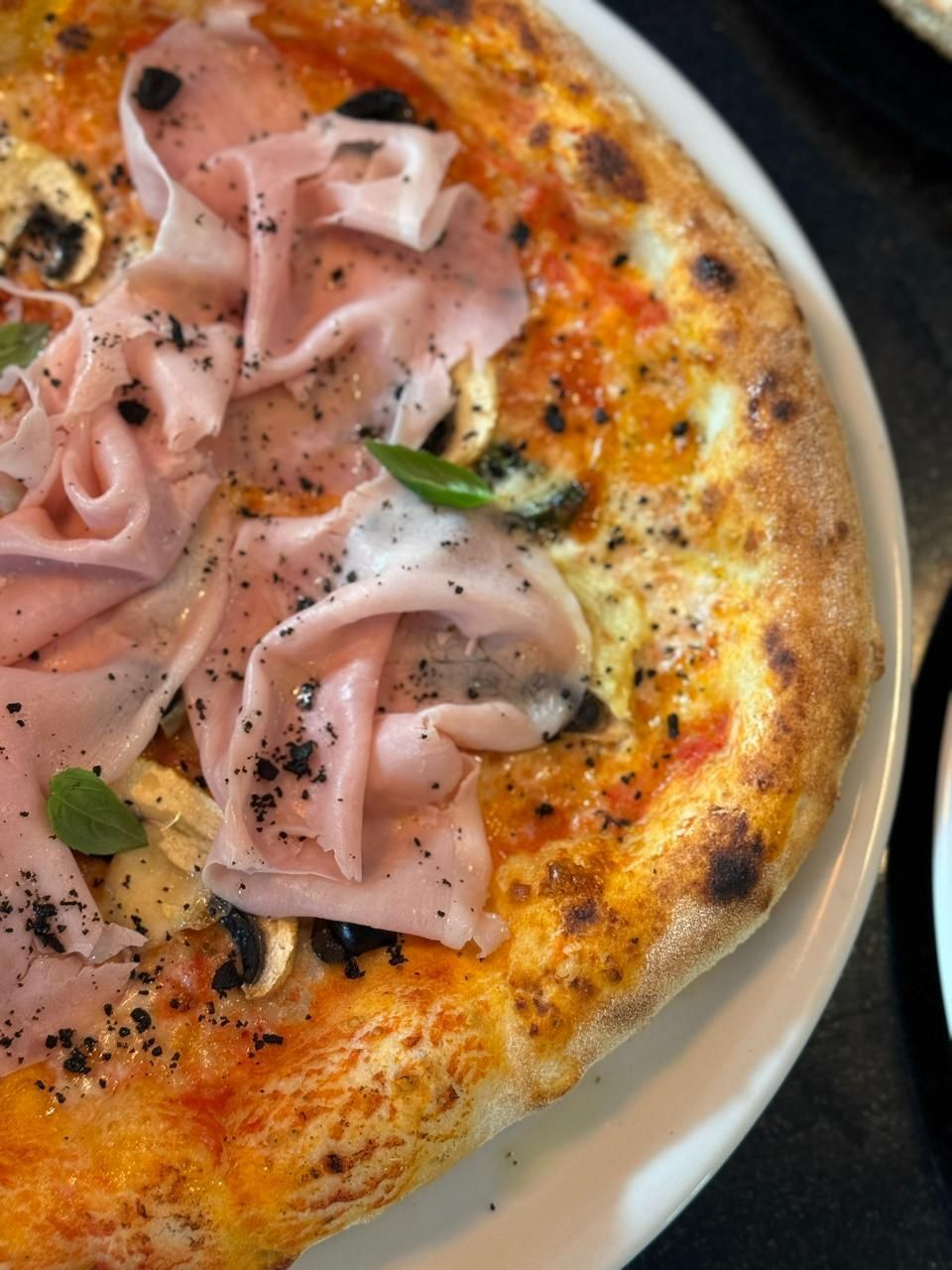 Pizza mit Schinken, Champignons und Kräutern auf einem weissen Teller; Nahaufnahme.
