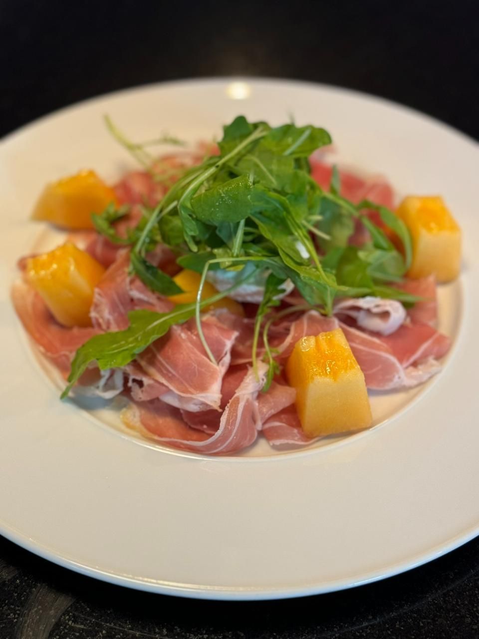 Prosciutto- und Cantaloupe-Salat mit Rucola auf weissem Teller.