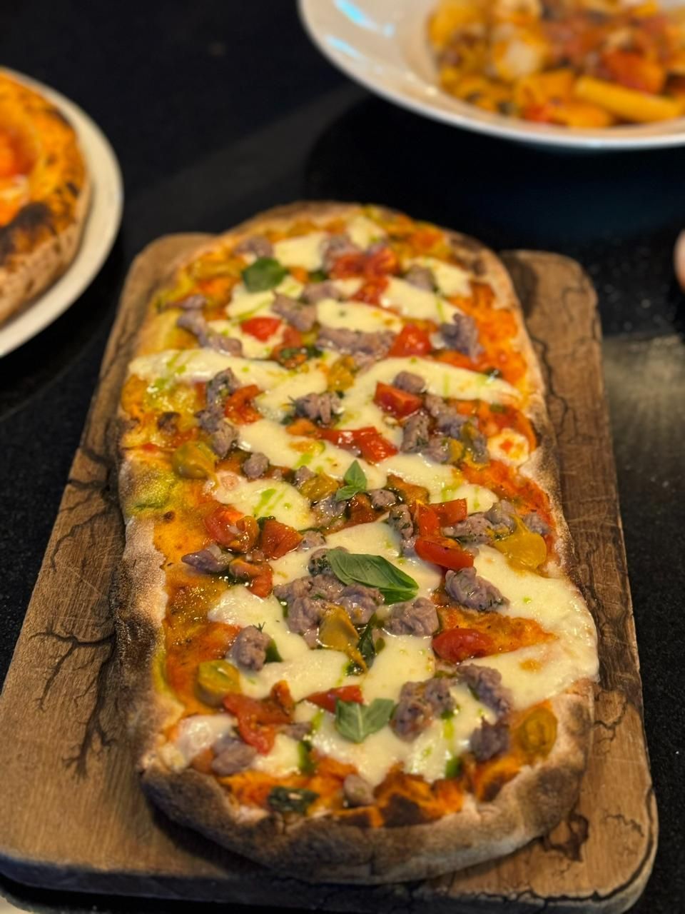 Rechteckige Pizza auf einem Holzbrett; Belag: Wurst, Tomaten, Basilikum und geschmolzener Käse.