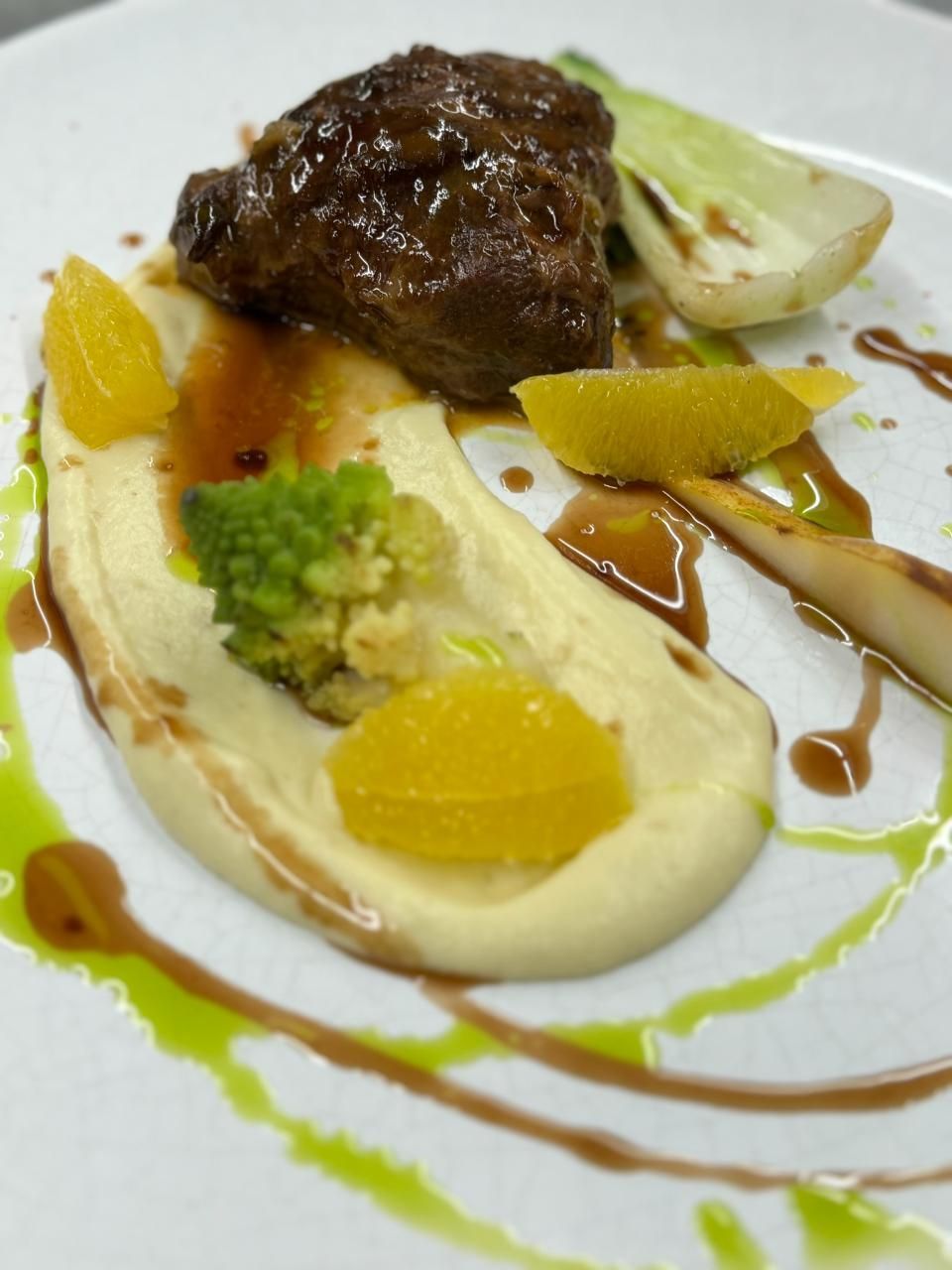 Geschmortes Fleisch auf cremigem Püree, garniert mit Orangenscheiben, grüner Sauce und Gemüse auf einem weissen Teller.
