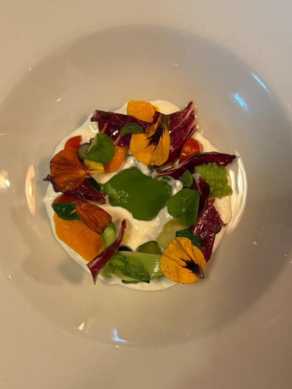 Weisser Teller mit cremefarbenem Boden, garniert mit buntem Gemüse, Kräutern und essbaren Blüten.