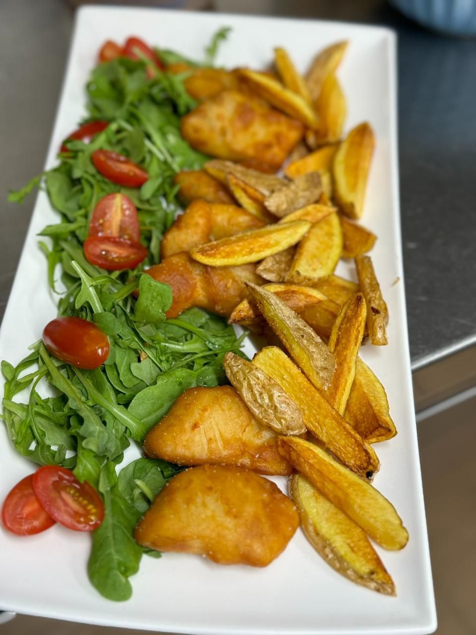 Gebratene Fischstücke, Kartoffelecken, Rucola und halbierte Kirschtomaten auf einer weissen Platte.