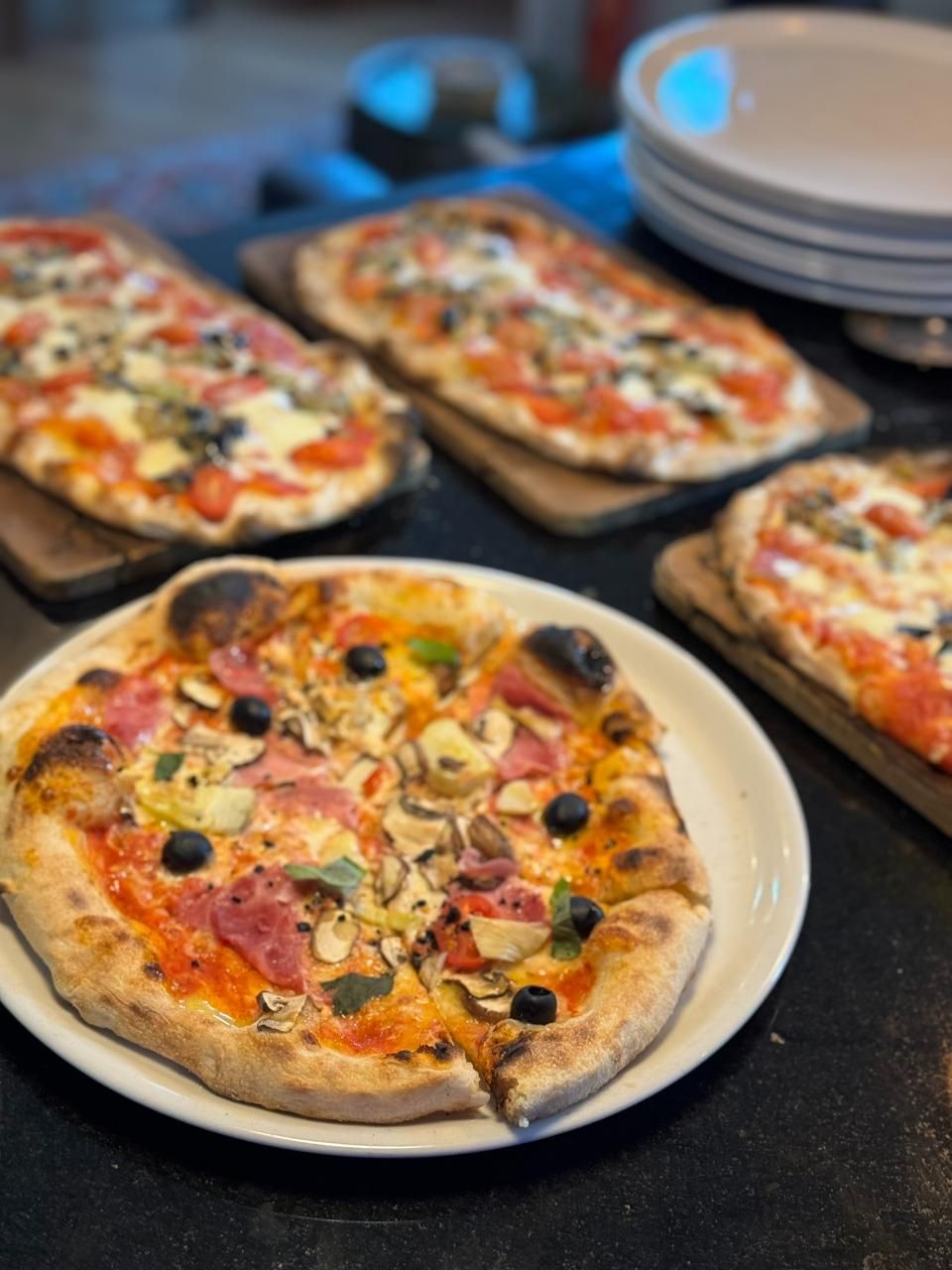 Vier Pizzen: eine runde auf einem Teller, drei rechteckige auf Holzbrettern. Belag: Tomaten, Champignons, Oliven.