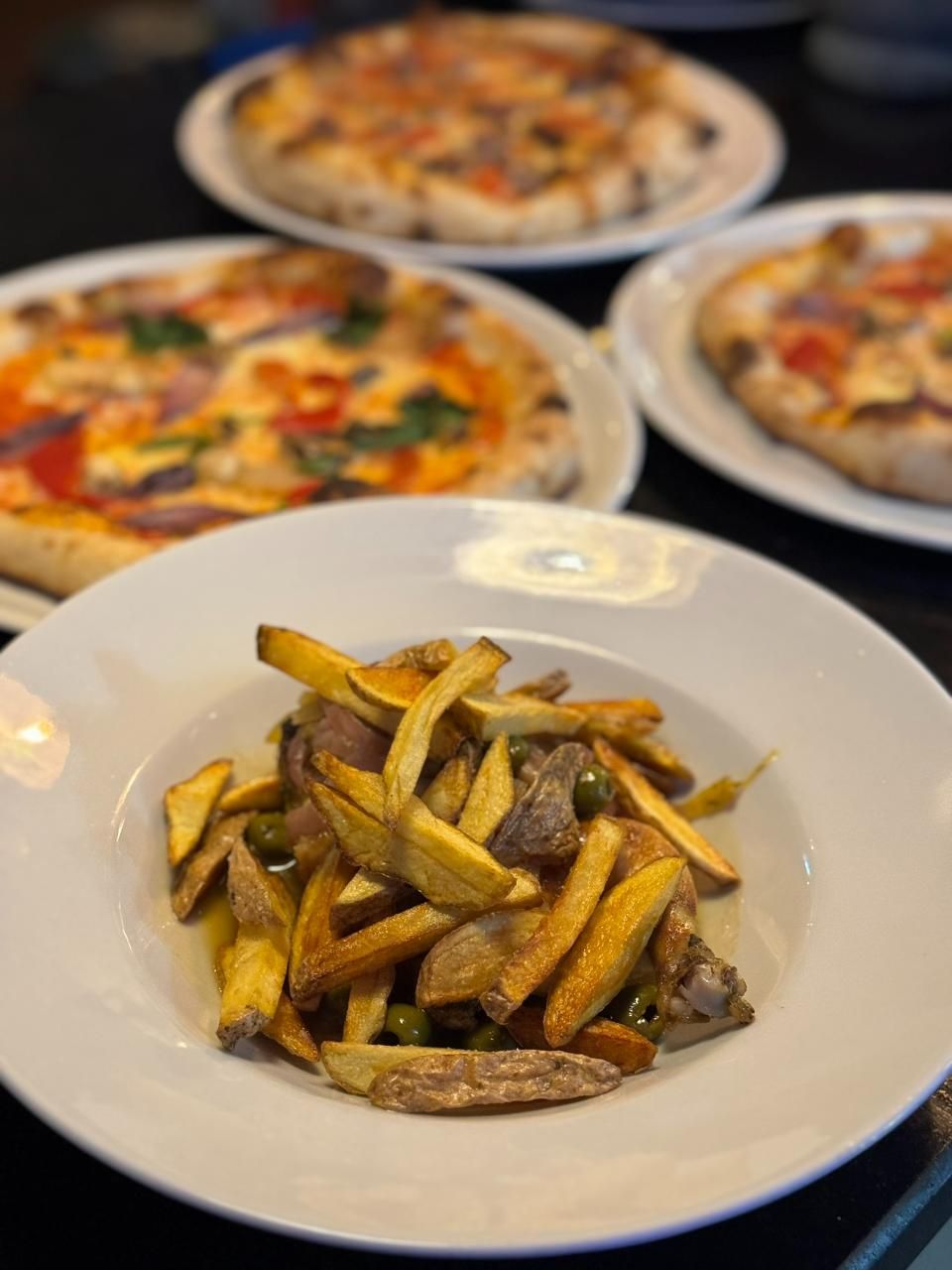 Pommes frites mit Fleisch und Erbsen in einer weissen Schüssel, im verschwommenen Hintergrund drei Pizzen.