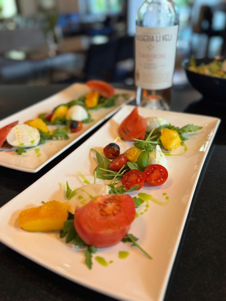 Caprese-Salat mit Tomaten, Mozzarella und Rucola auf rechteckigen weissen Tellern. Im Hintergrund eine Weinflasche.
