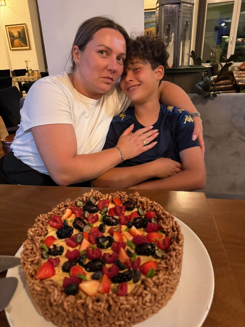 Frau und Junge umarmen sich lächelnd; auf einem Tisch steht ein Obstkuchen. Szene in einem Restaurant.