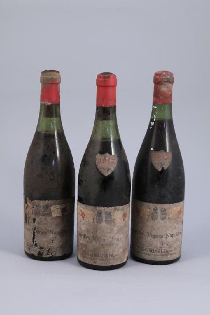 Trois vieilles bouteilles de vin aux étiquettes usées et aux capsules rouges, dressées sur un fond uni.