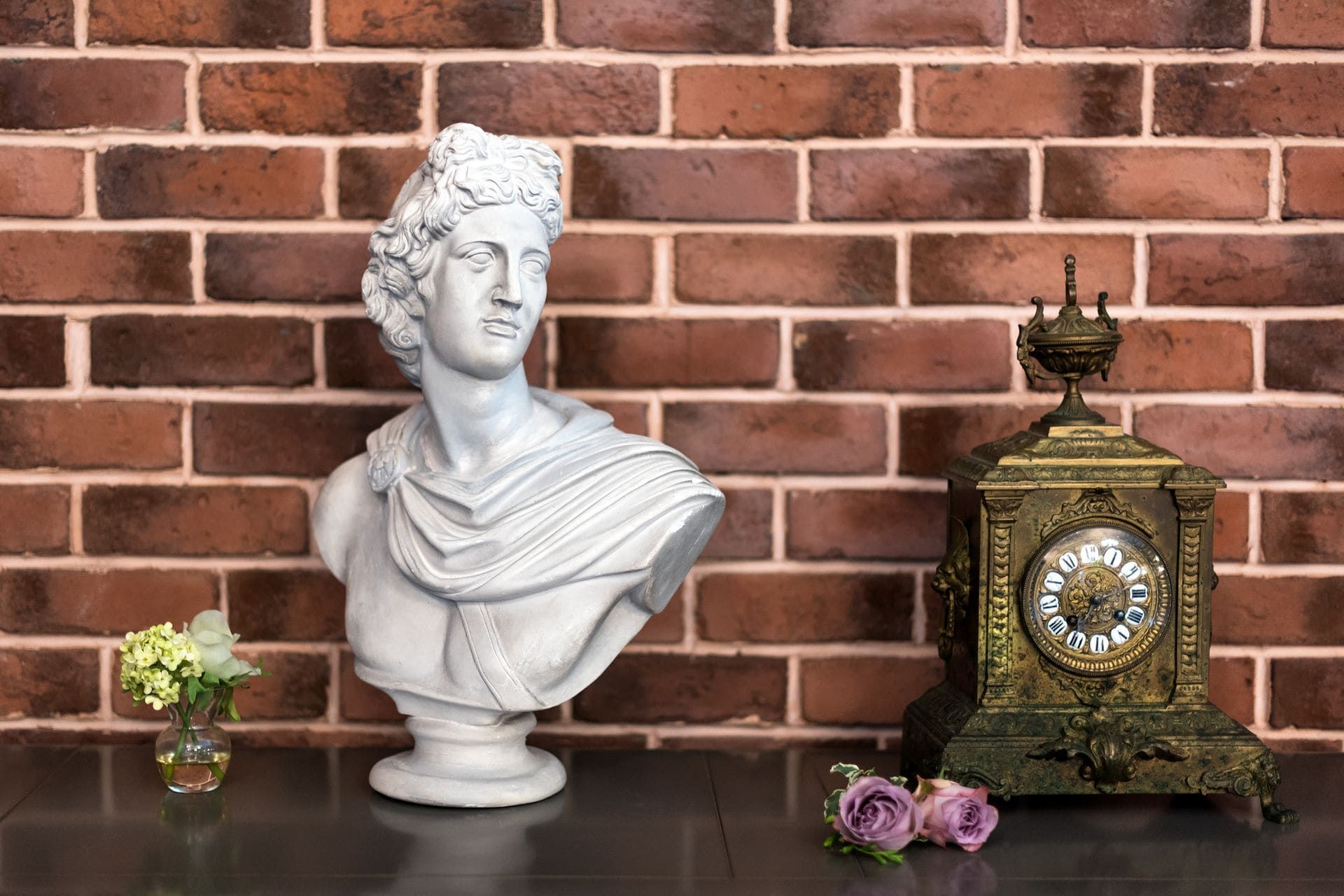 Buste blanc, horloge ornée et petites fleurs sur une table noire, devant un mur de briques
