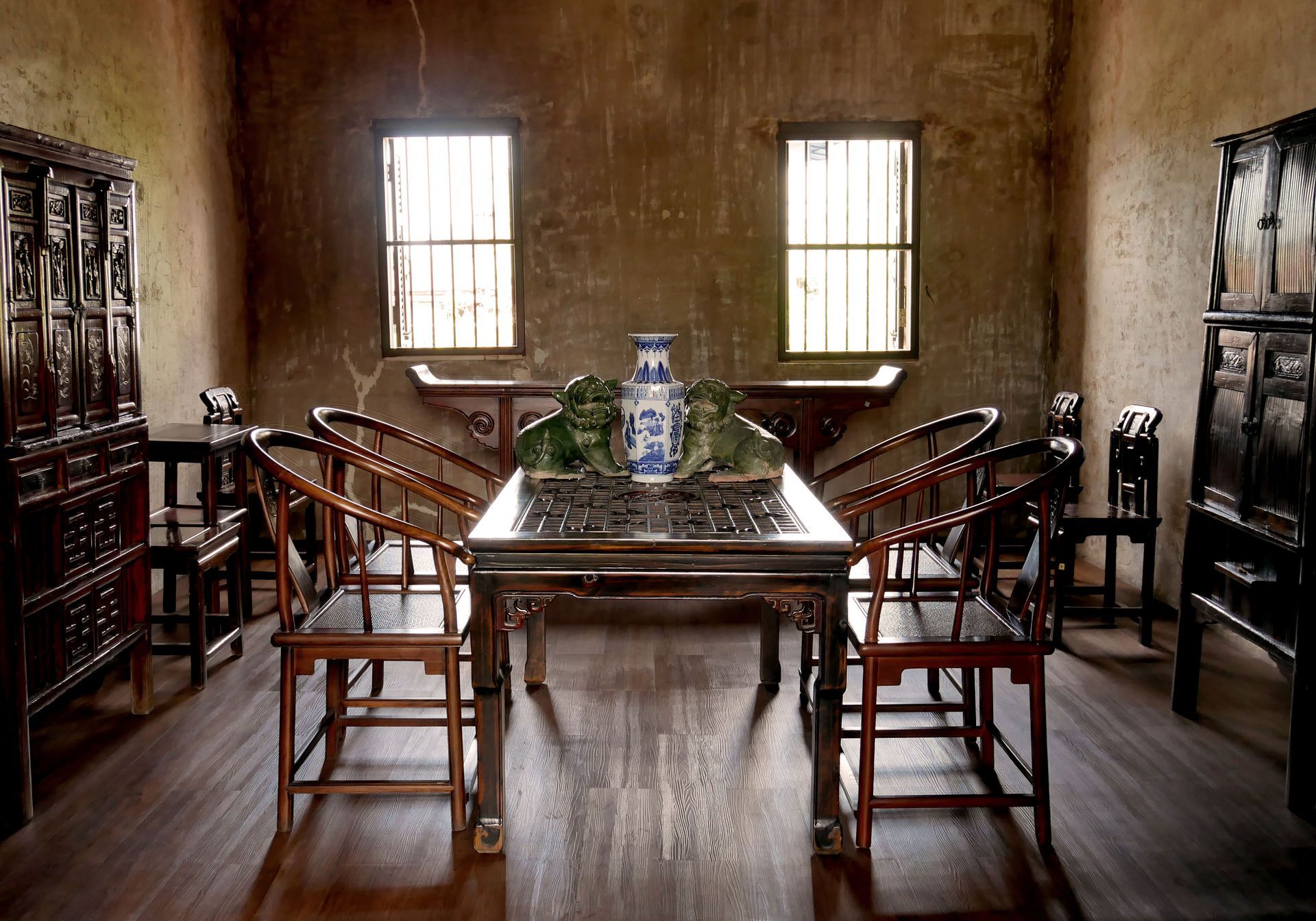 Salle à manger rustique avec table en bois, chaises, armoires et deux fenêtres avec voilages