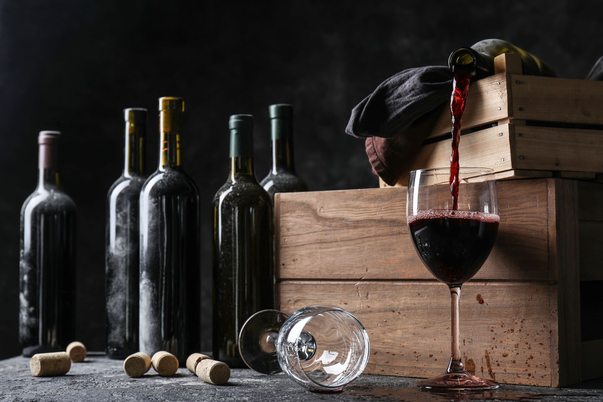Des bouteilles de vin et un verre de vin rouge à côté de caisses en bois empilées et de bouchons de liège sur un fond sombre.