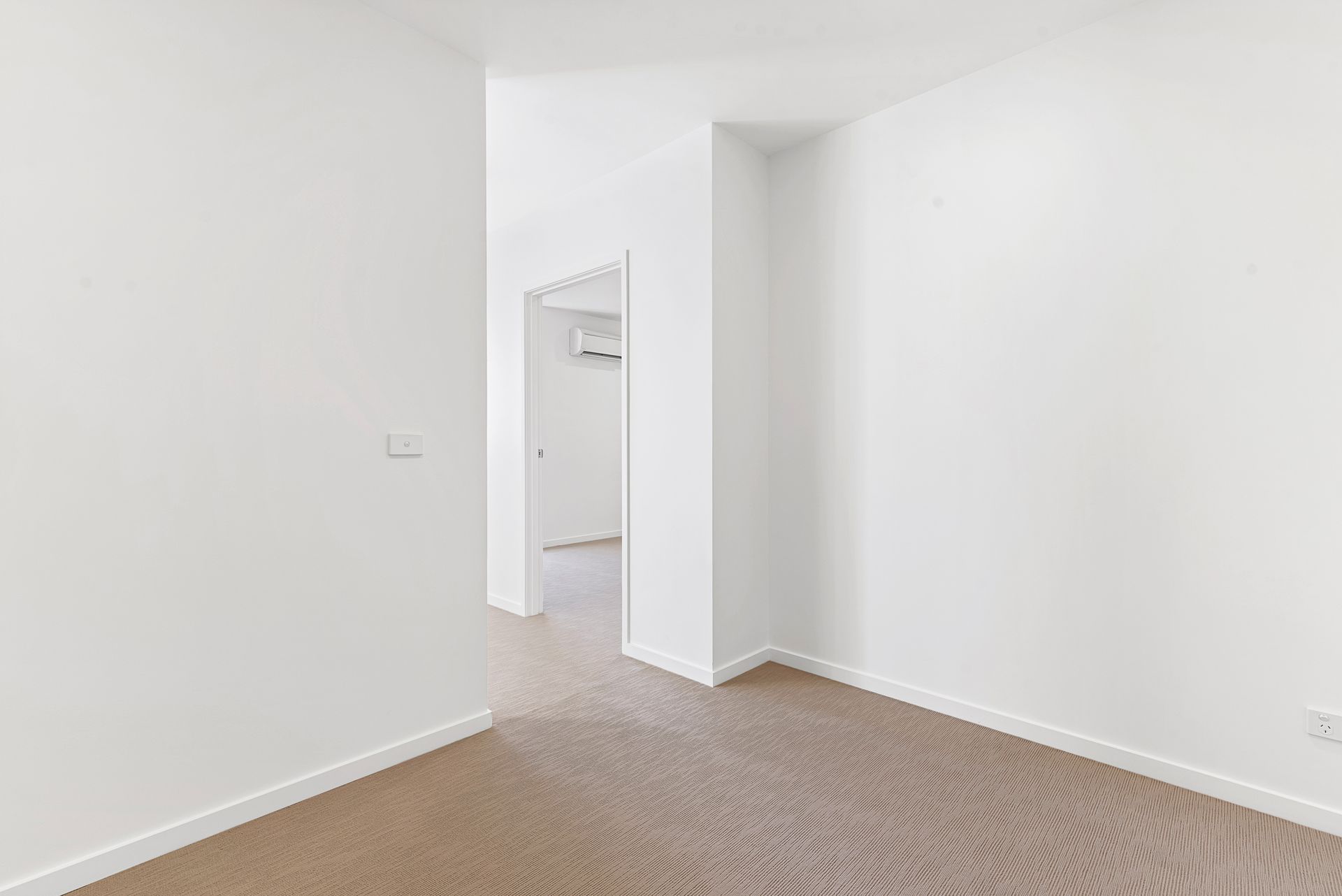 Pièce blanche vide avec moquette beige et porte ouverte donnant sur un hall