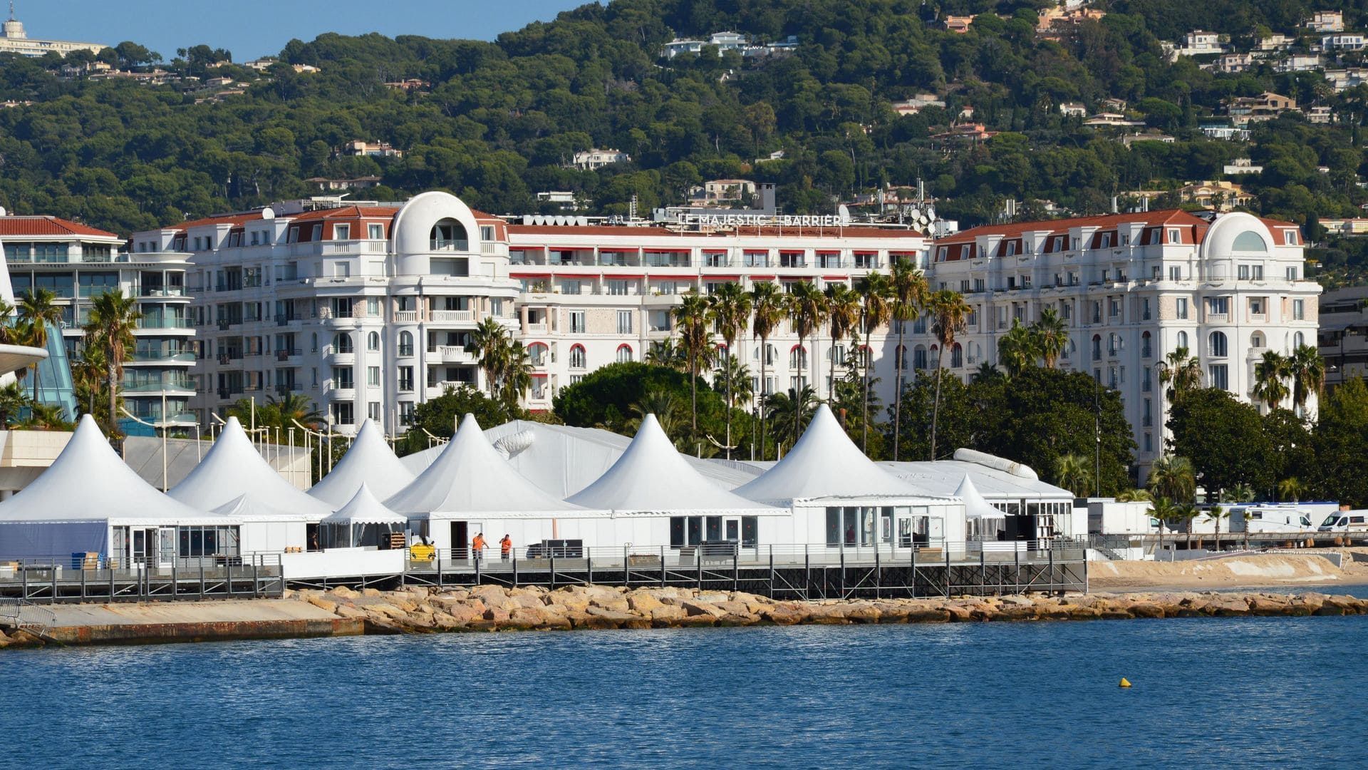 Hôtel blanc en bord de mer avec des toits rouges et des tentes blanches le long d'un rivage bleu, adossé à des collines verdoyantes