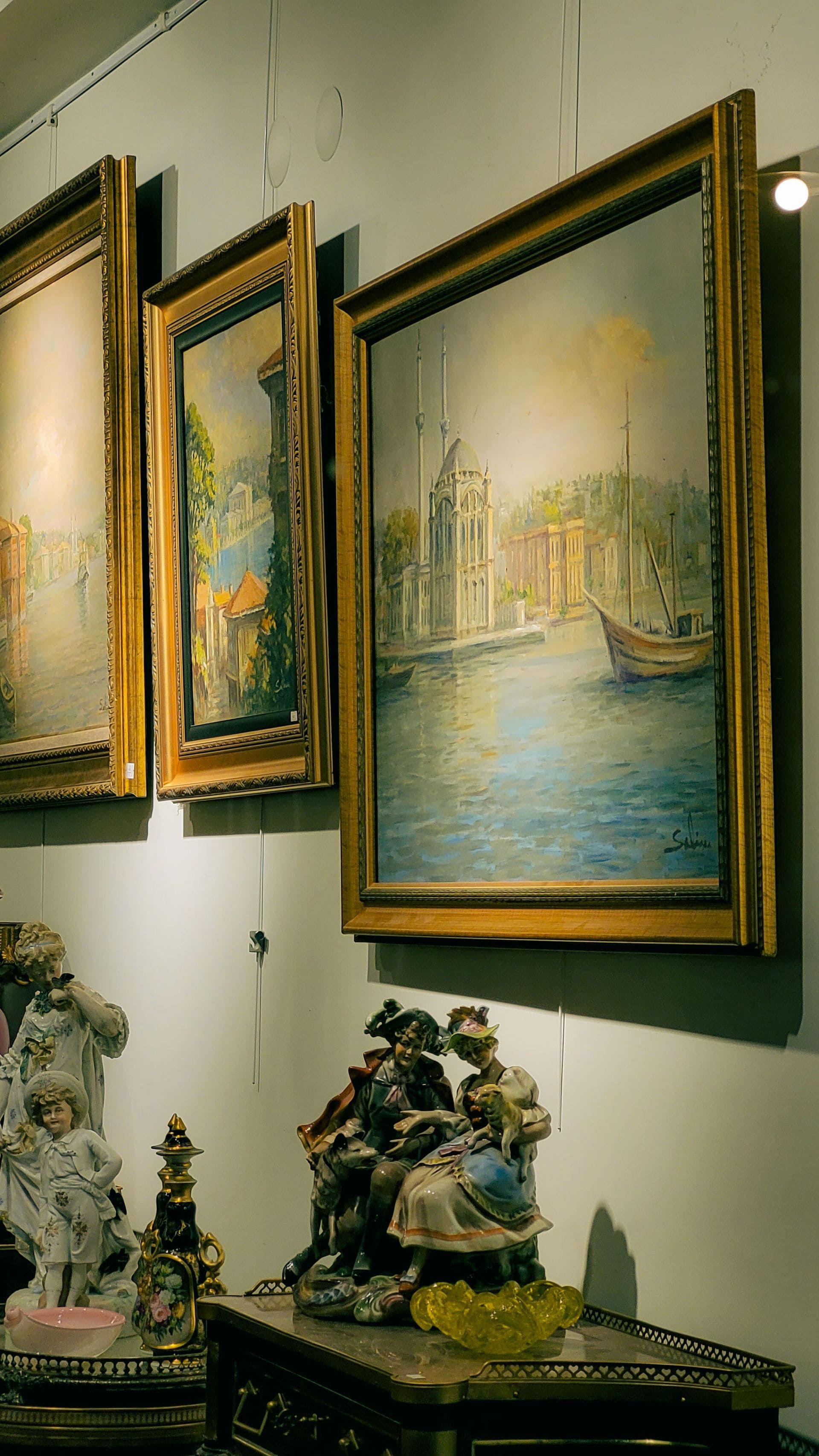 Des tableaux encadrés représentant des scènes vénitiennes sont accrochés au mur d'une galerie, au-dessus de sculptures décoratives et d'une petite table.