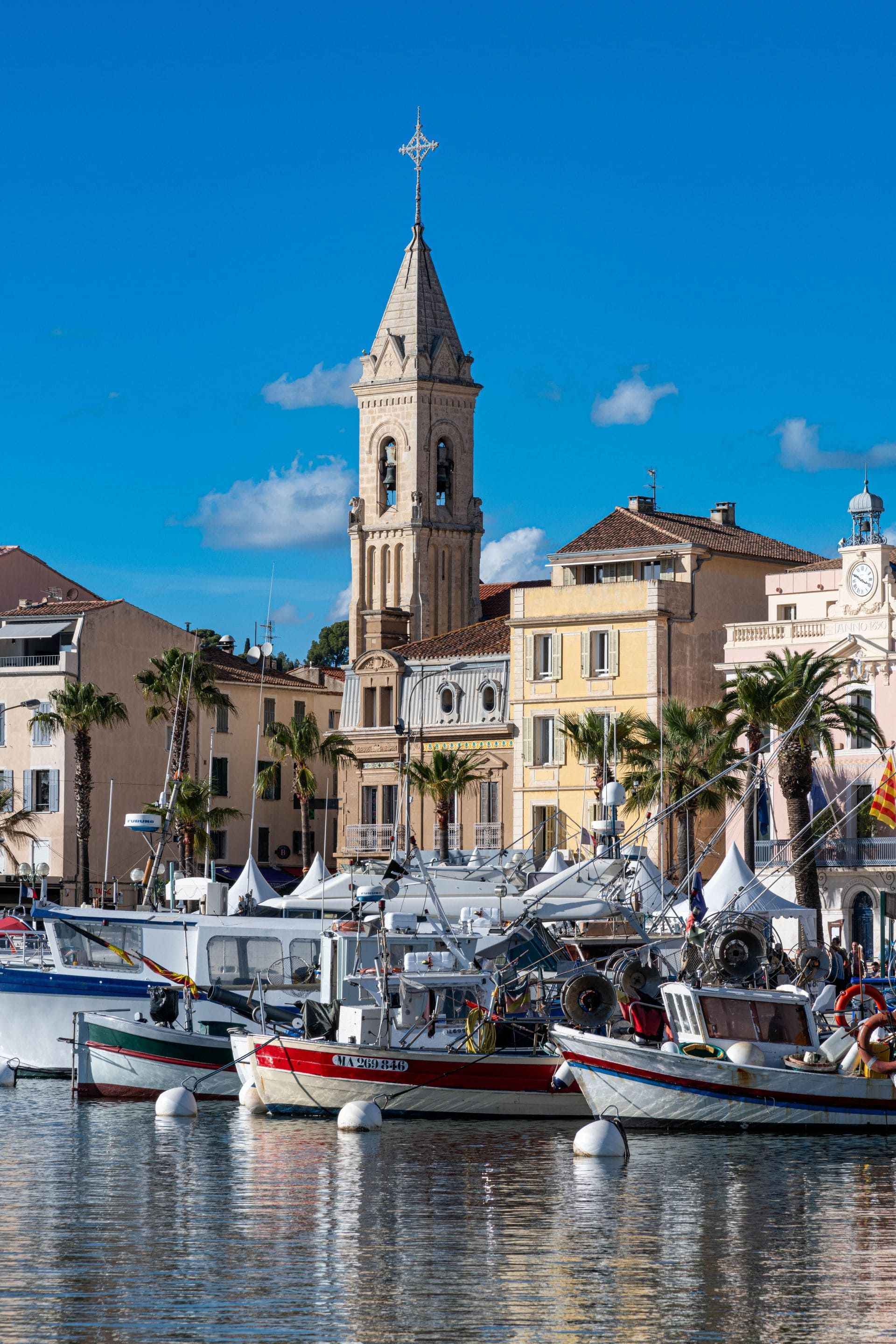 Un port avec des bateaux à quai, des palmiers et un clocher historique sous un ciel d'un bleu éclatant