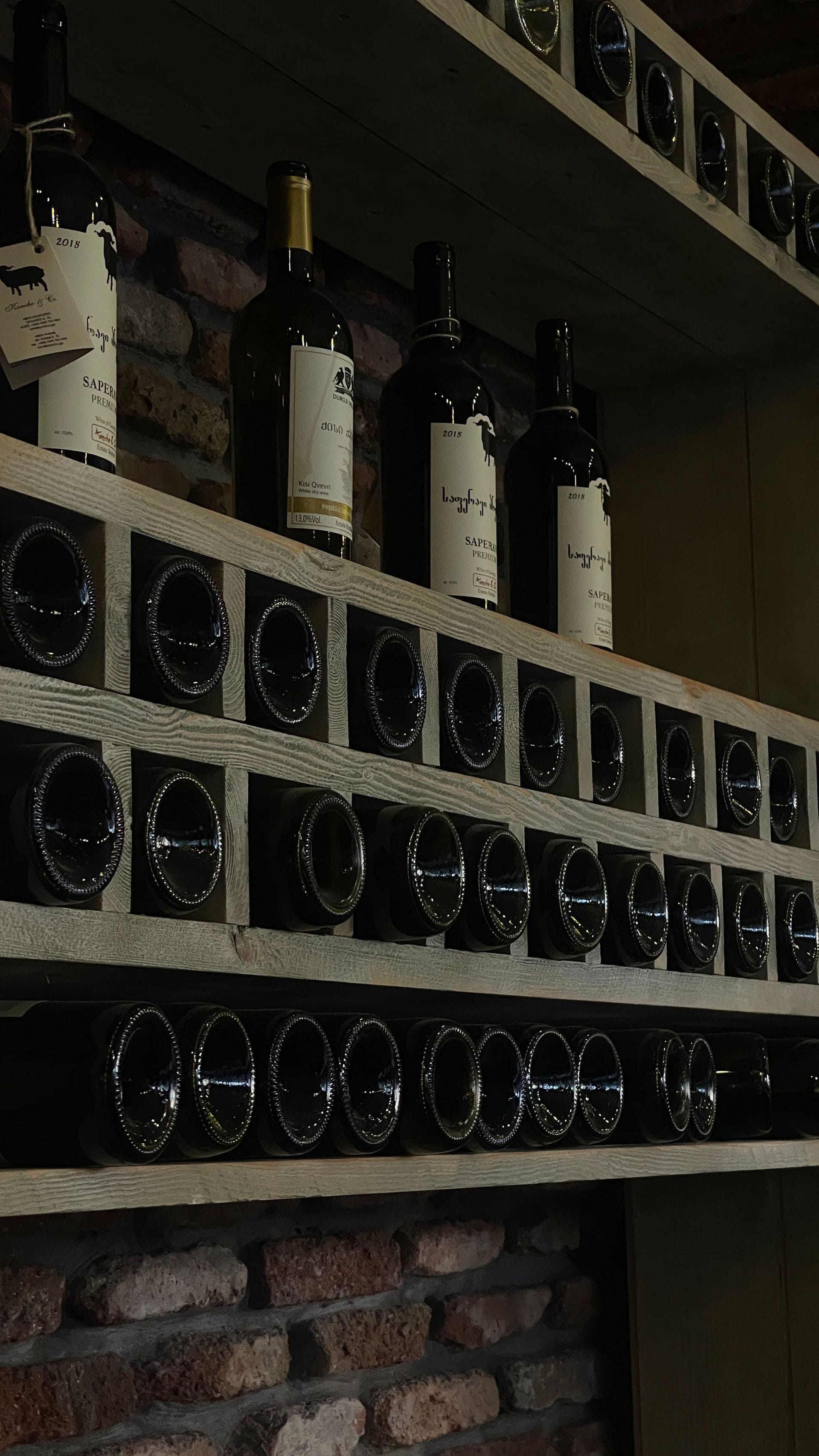 Des bouteilles de vin sont exposées sur une étagère murale rustique, au-dessus de casiers à vin empilés.