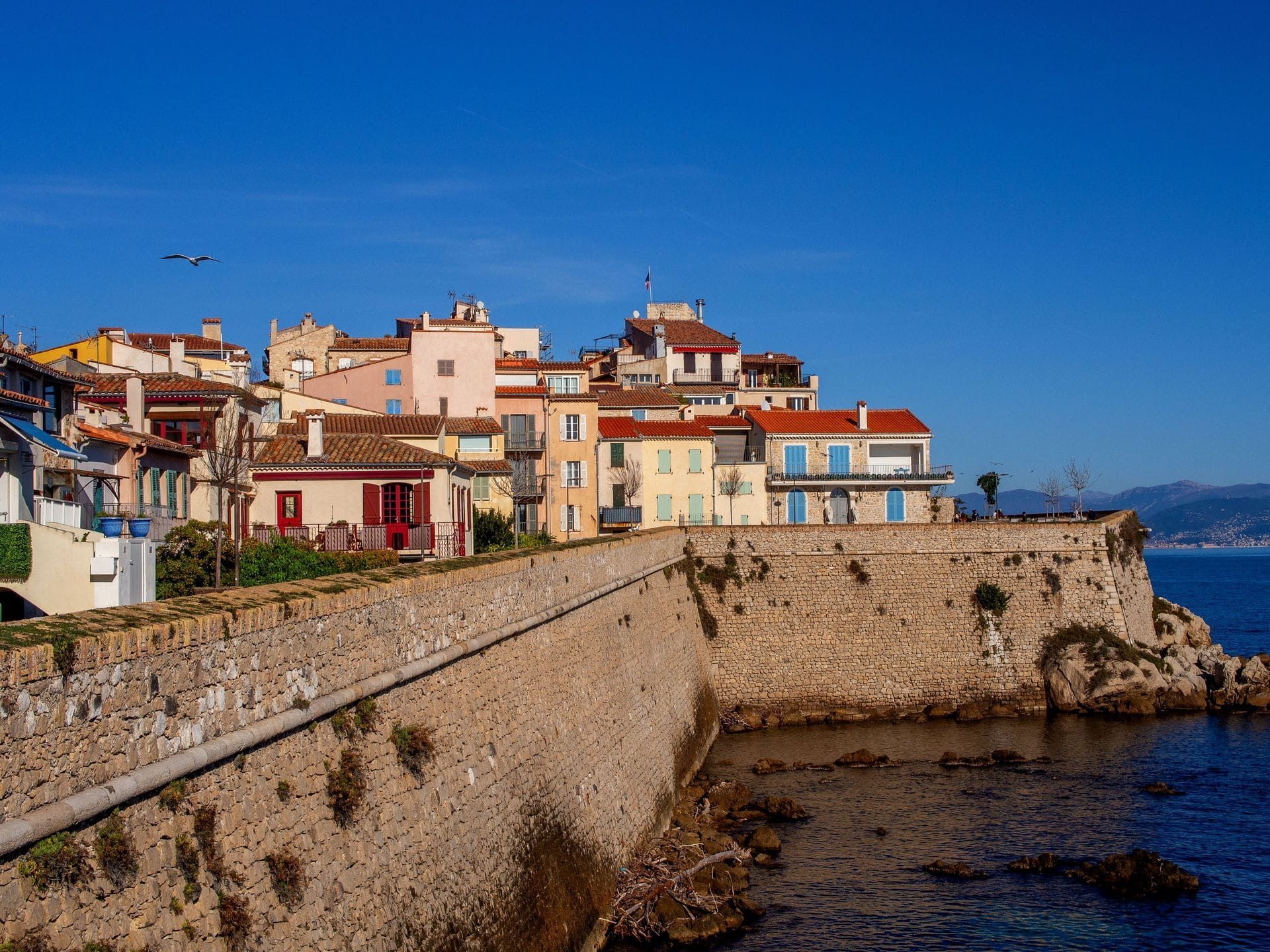 Village côtier à flanc de falaise, avec ses maisons colorées et son mur de pierre surplombant la côte rocheuse sous un ciel bleu