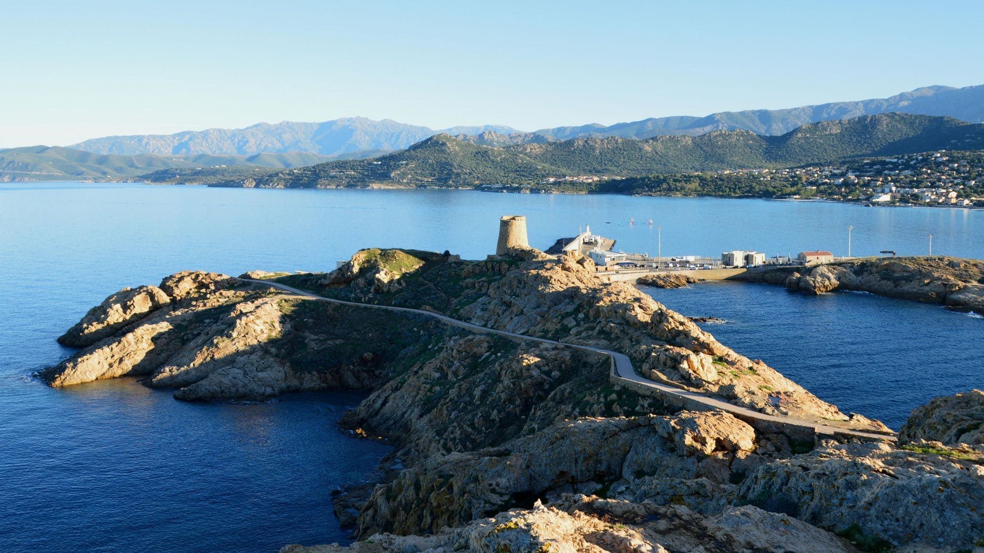 Île rocheuse avec une petite tour en pierre, entourée par la mer bleue et des montagnes lointaines sous un ciel clair