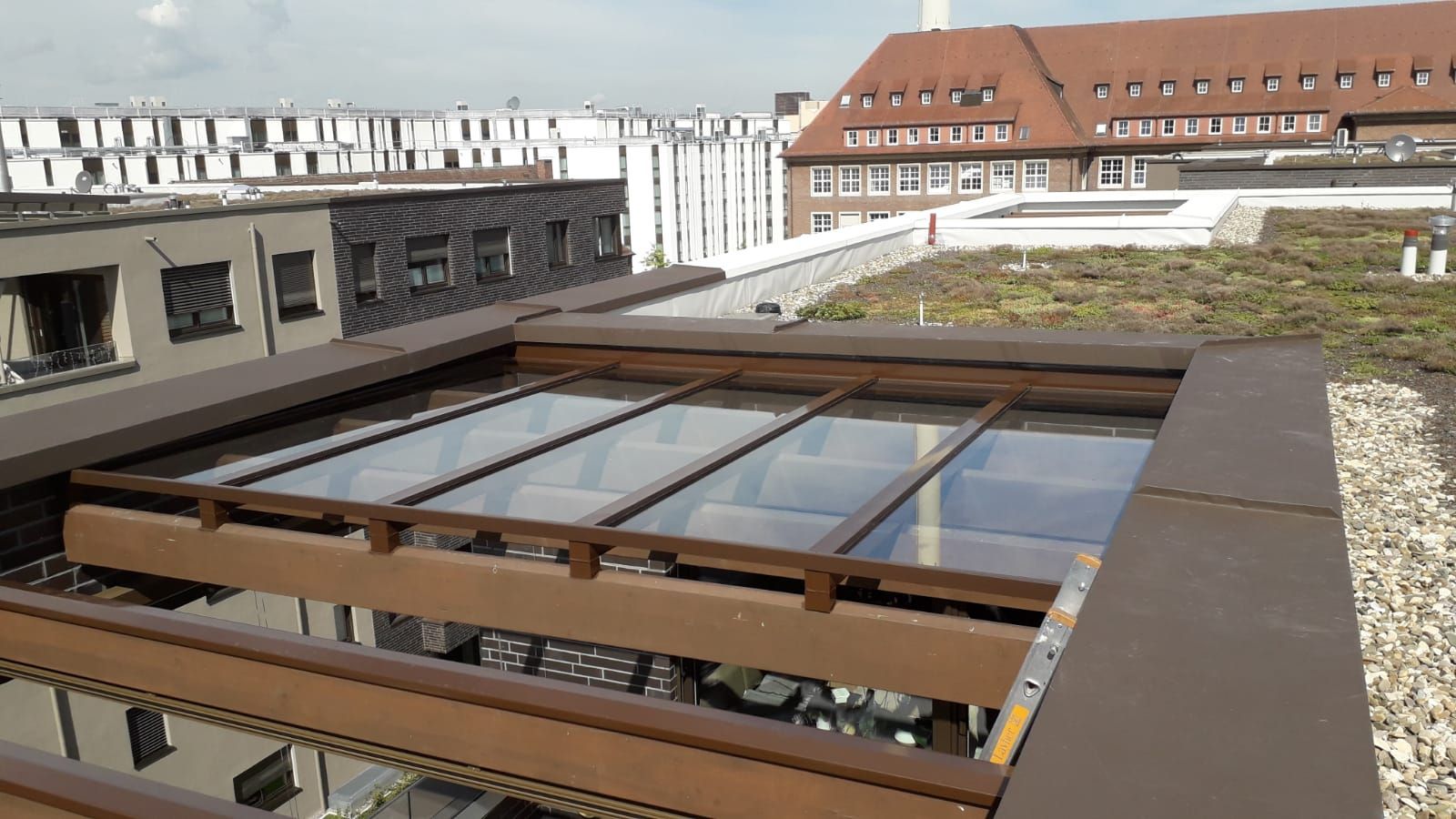 Dach mit Glasplatten, braunen Rahmen. Grünes Dach und Gebäude im Hintergrund. Sonniger Tag.