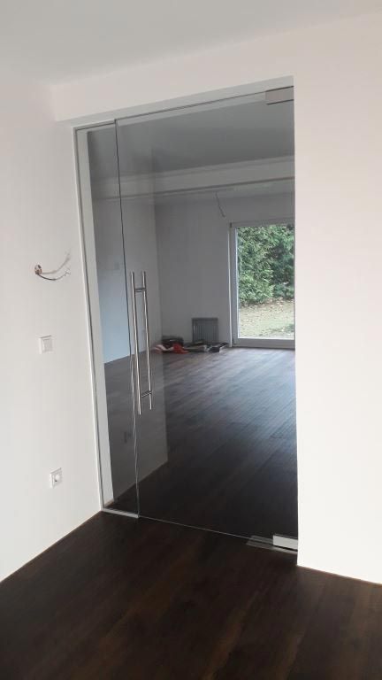 Glastür in einem Raum mit weißen Wänden und dunklem Holzboden, die zu einem anderen Raum mit einem Fenster führt.