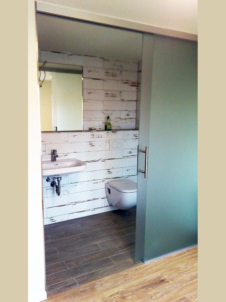 Badezimmer mit Milchglas-Schiebetür, weißer, verwitterter Holzwand, Toilette, Waschbecken und Boden in Holzoptik.