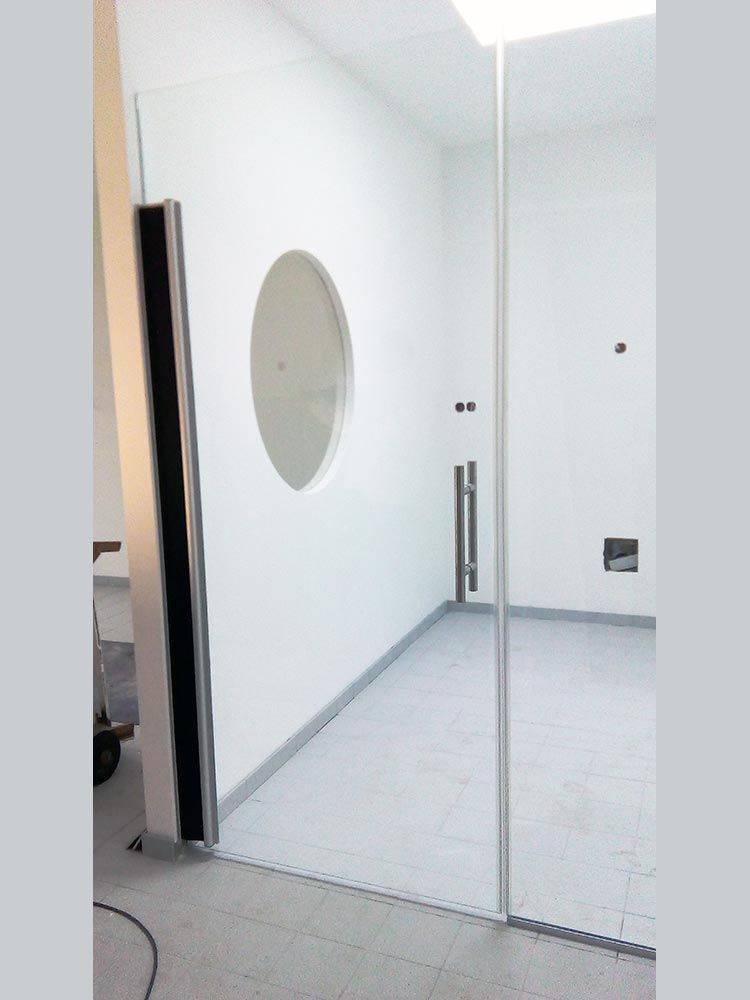 Eine Glastür mit Bullauge öffnet sich zu einem weißen Raum mit Edelstahlgriff und grauem Boden.