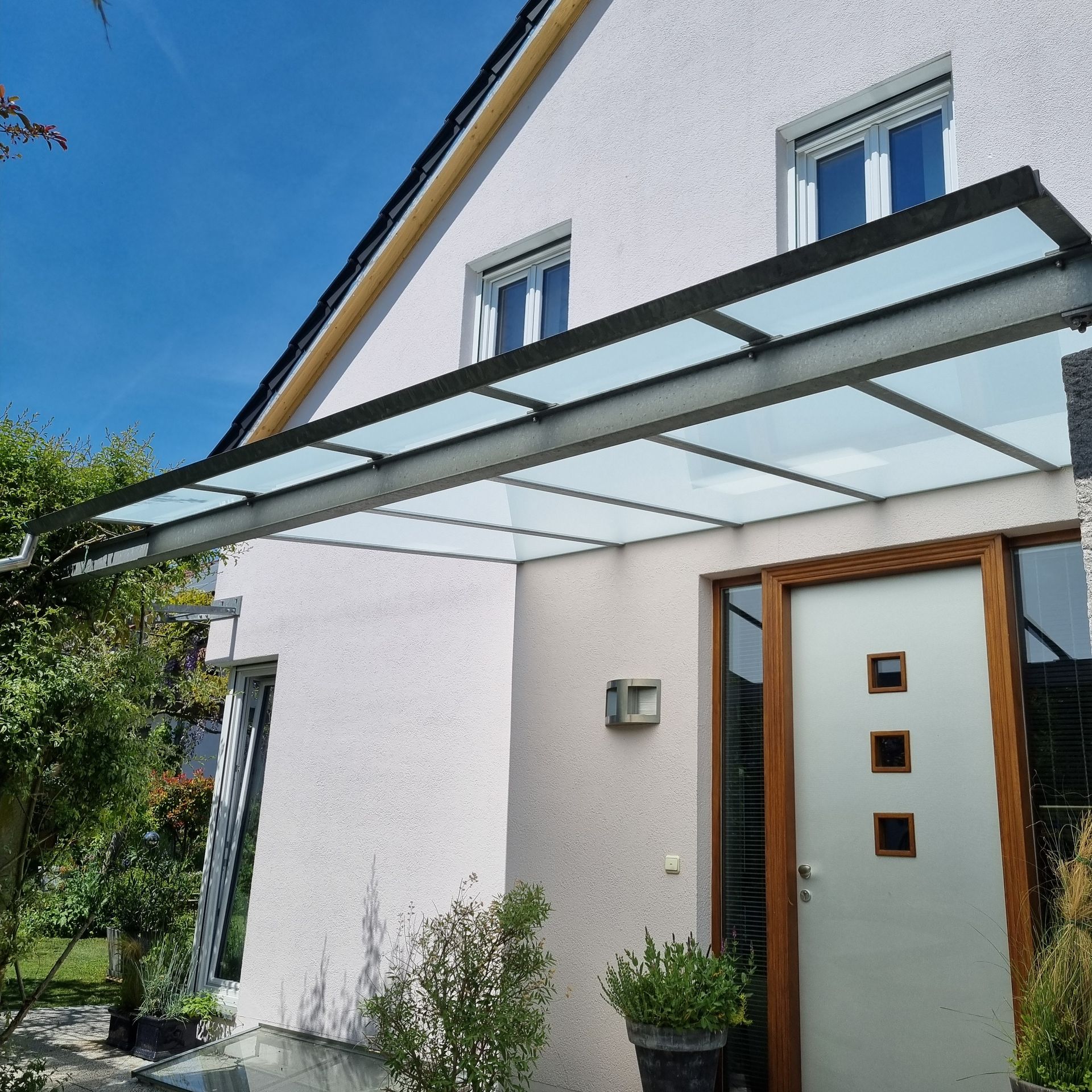 Moderner Hauseingang mit Glasmarkise und weißen Wänden. Braune Tür mit drei quadratischen Fenstern. Blauer Himmel.