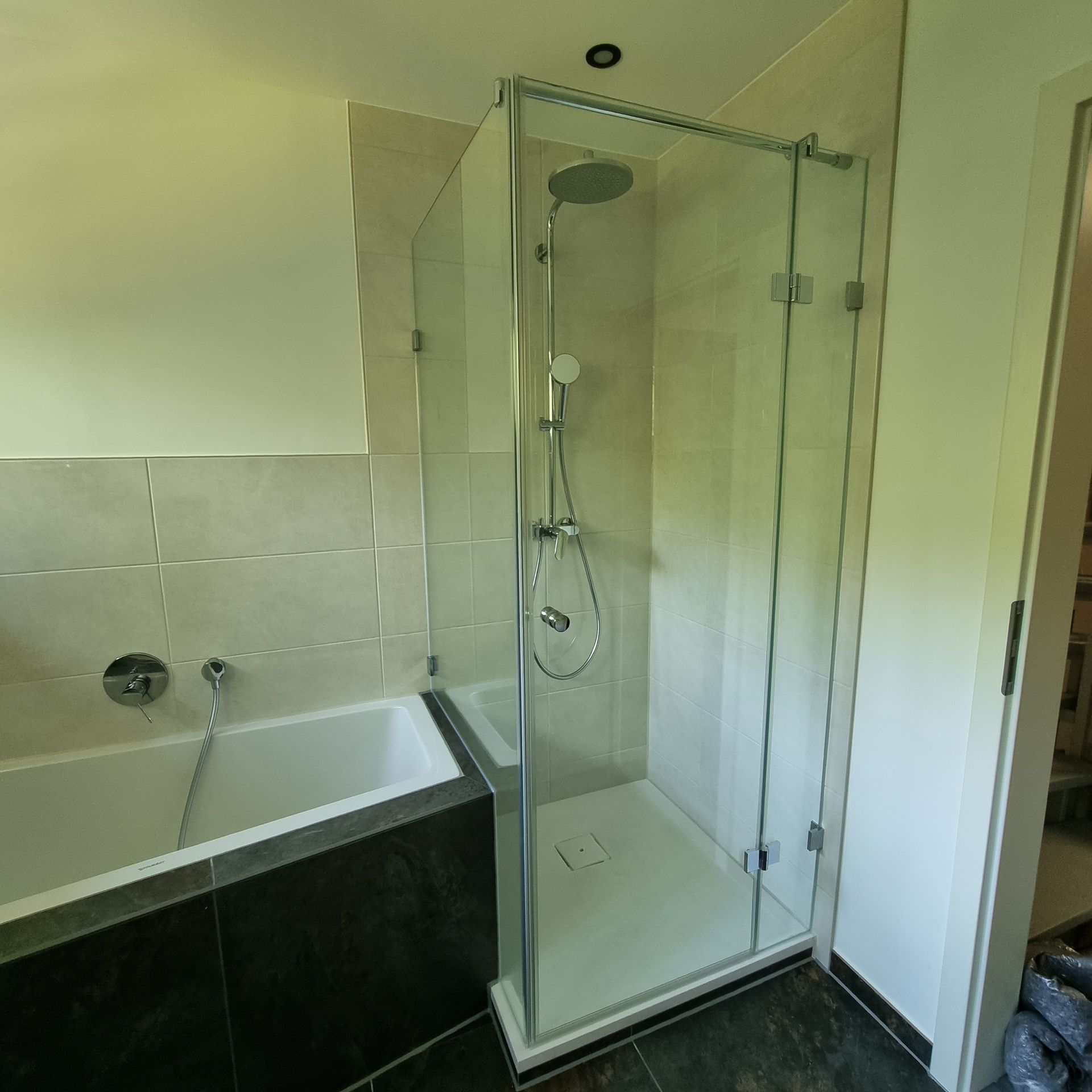 Badezimmer mit weißer Badewanne, grauen Fliesen und einer Dusche aus klarem Glas mit Chromarmaturen.