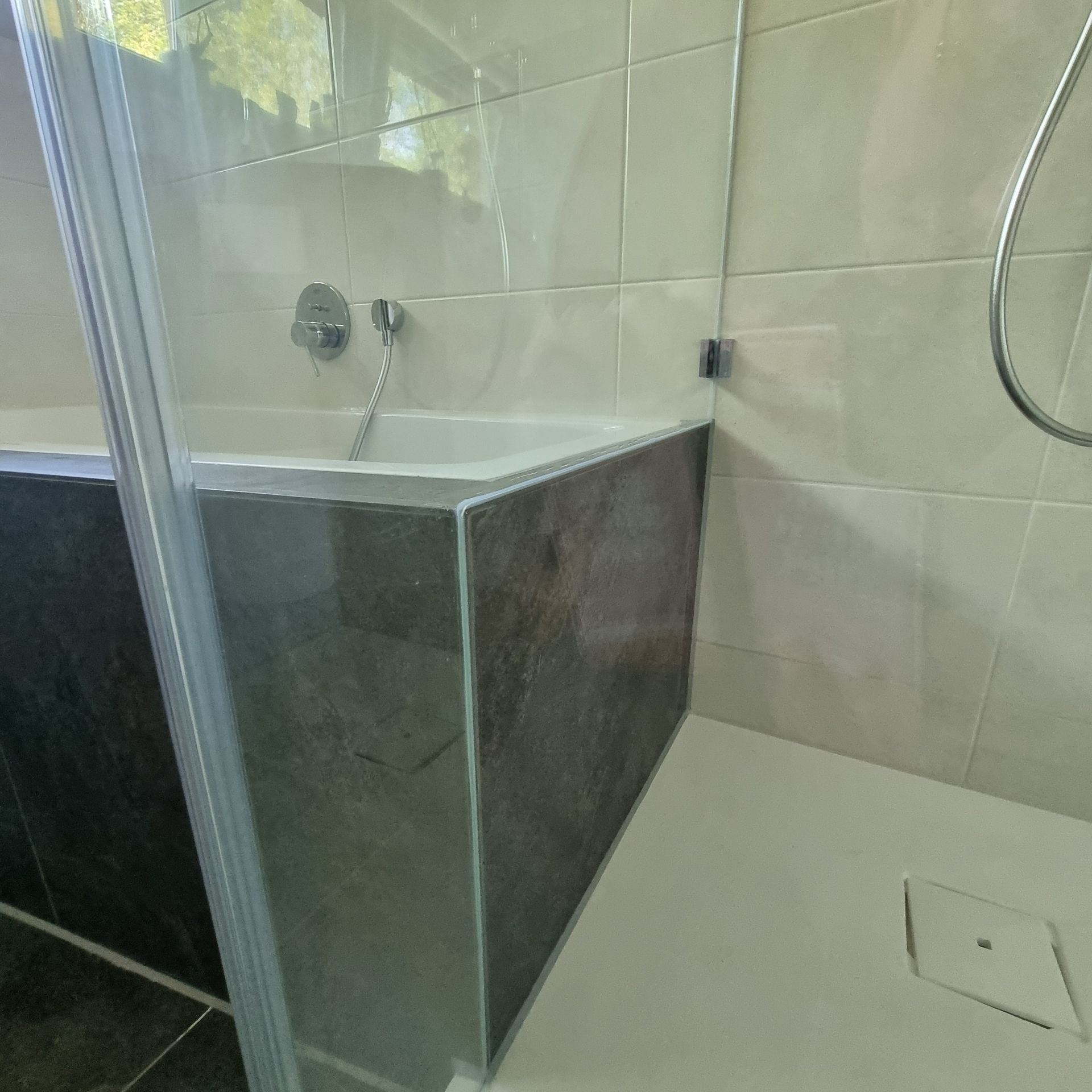 Badezimmer mit Badewanne und Dusche. Dunkelgraue Fliesen an der Wannenseite. Weißer Duschboden mit Abfluss.