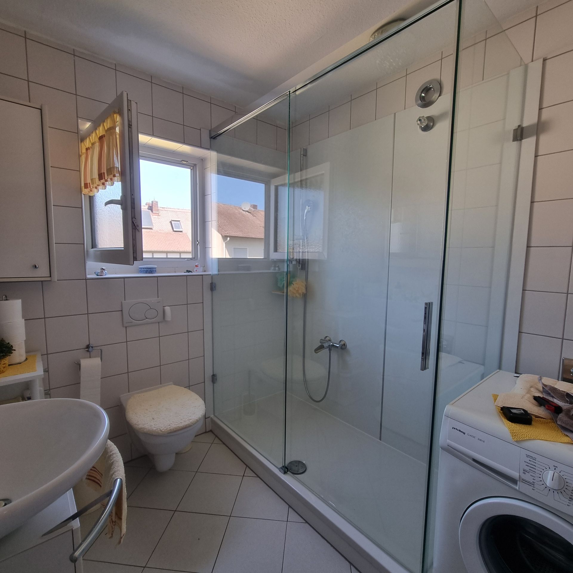 Badezimmer mit Glasdusche, Toilette, Waschbecken und Waschmaschine. Es gibt ein kleines offenes Fenster mit Aussicht.