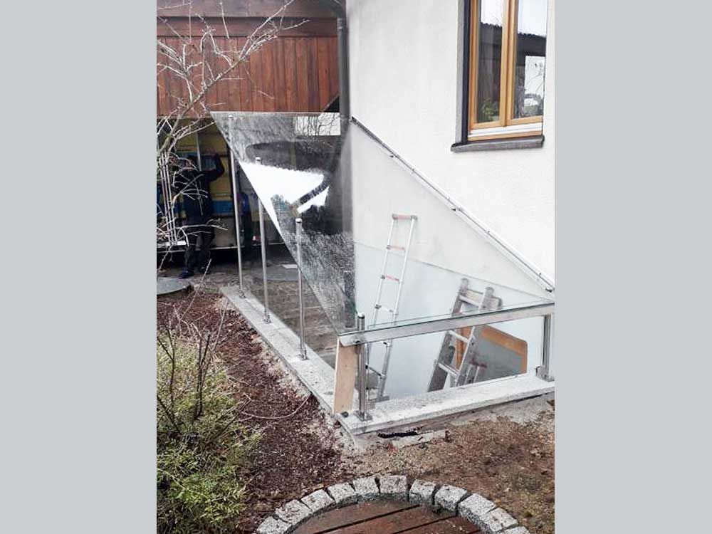 Verglaster Kellereingang mit Glasgeländer vor einem weißen Gebäude. Im Inneren steht eine Leiter.