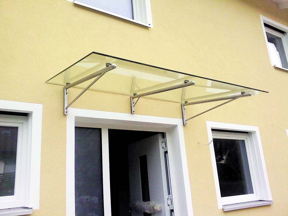Glasmarkise über einer weißen Tür an einem gelben Gebäude, getragen von silbernen Klammern.