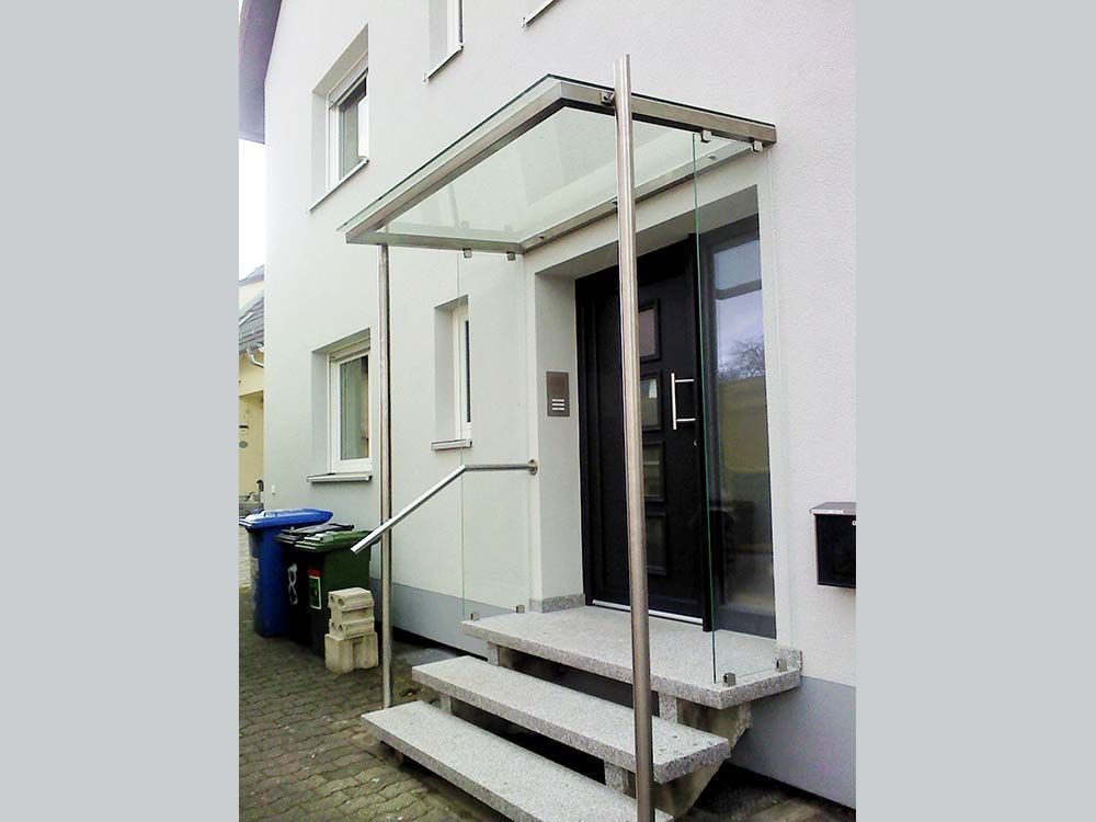 Außenansicht eines weißen Gebäudes mit einem Vordach aus Metall und Glas über dem Eingang und den Stufen.