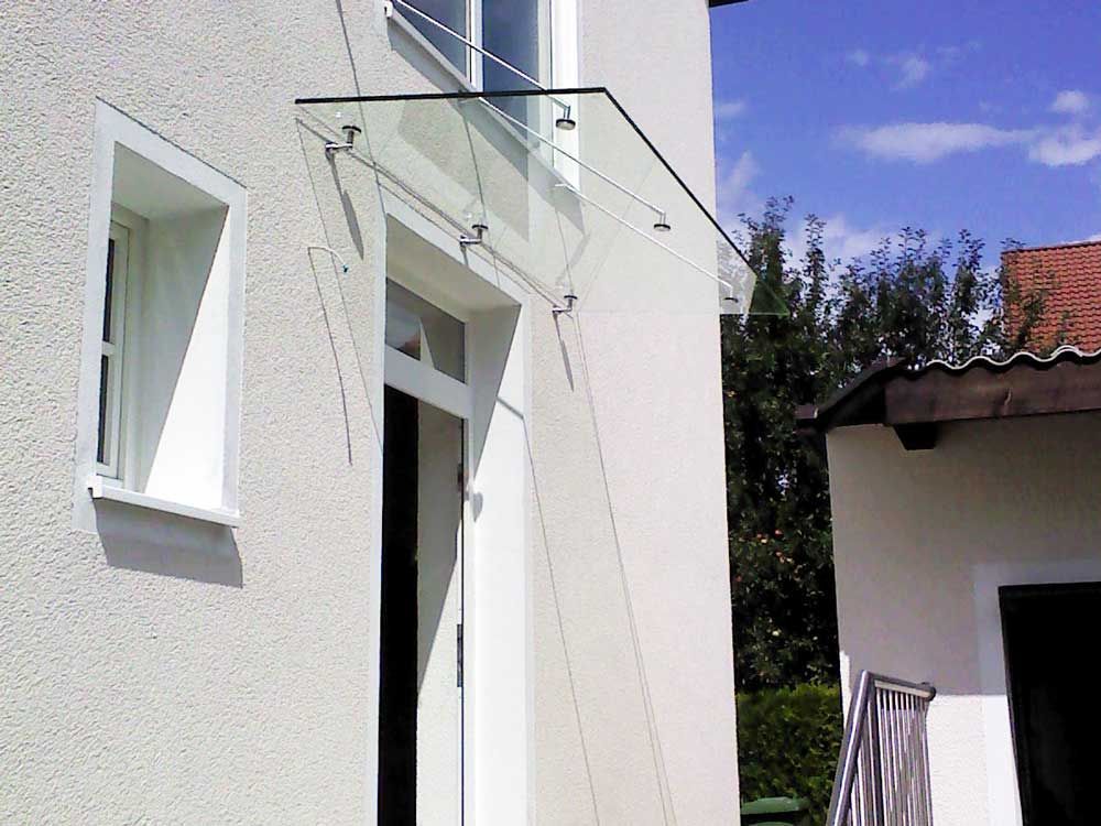 Glasmarkise über einer weißen Tür, mit einem kleinen Fenster links, vor einer weißen Stuckwand.