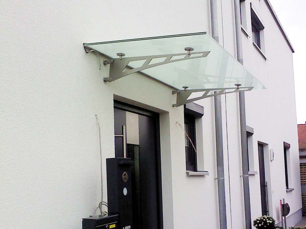Glasvordach über einer dunklen Tür an einem weißen Haus. Edelstahlstützen, trübes Glas.