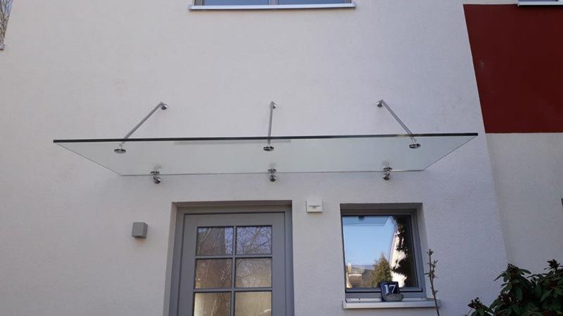 Eine Glasmarkise über einer Haustür und einem Fenster an einem hellen Gebäude.