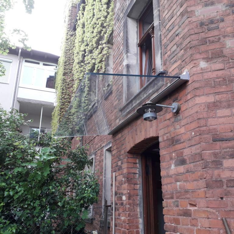 Backsteingebäude mit überdachtem Eingang und Fenster, an dessen Seite Efeu hochwächst. Eine Wandleuchte ist vorhanden.