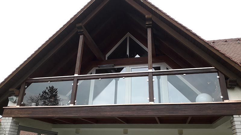 Brauner Holzbalkon mit Glaspaneelen an einem Haus mit Spitzdach.
