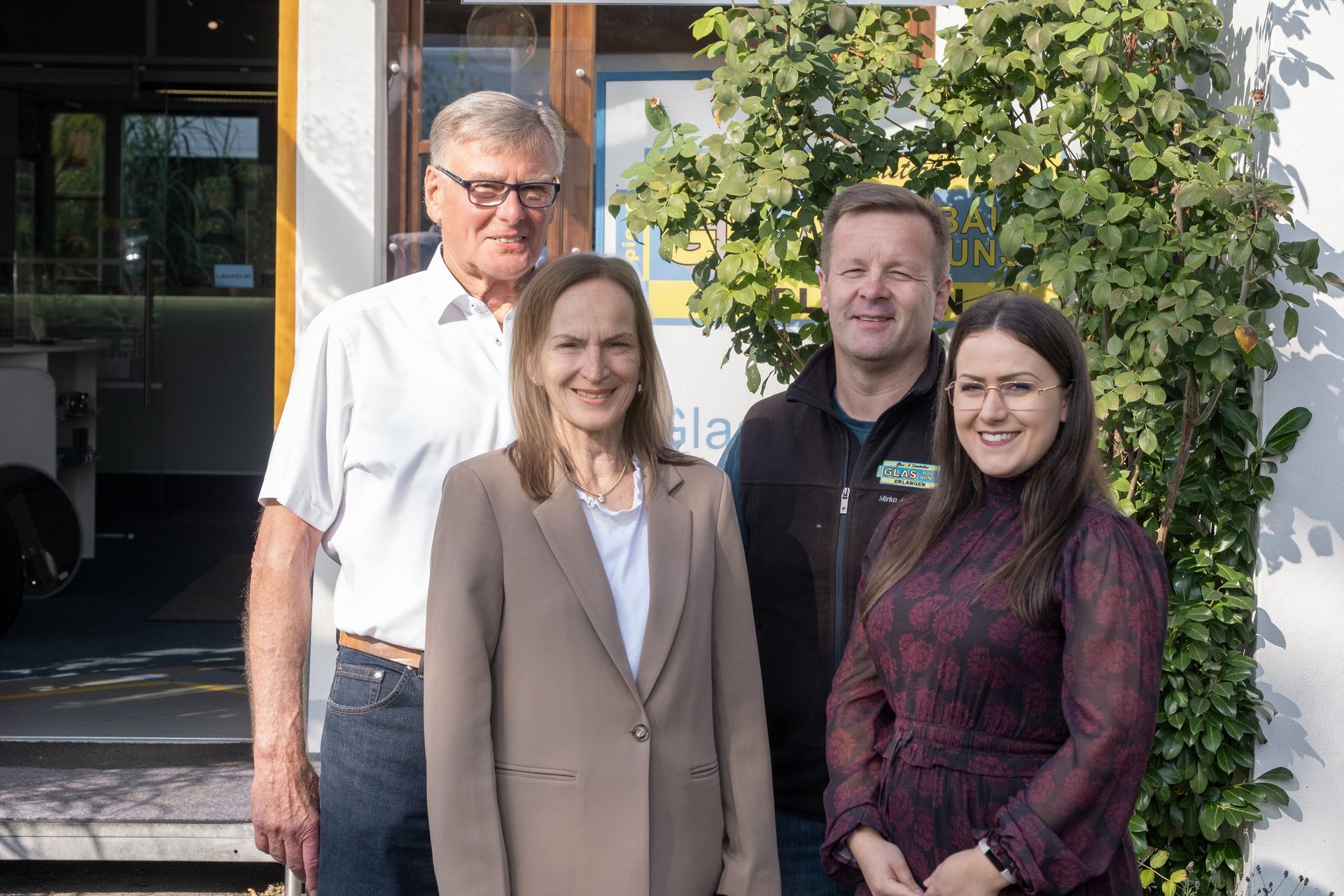Vier Personen posieren vor einem Gebäude. Zwei Frauen und zwei Männer stehen dicht beieinander und lächeln. Die rechte Seite des Gebäudes ist mit Laub bedeckt.