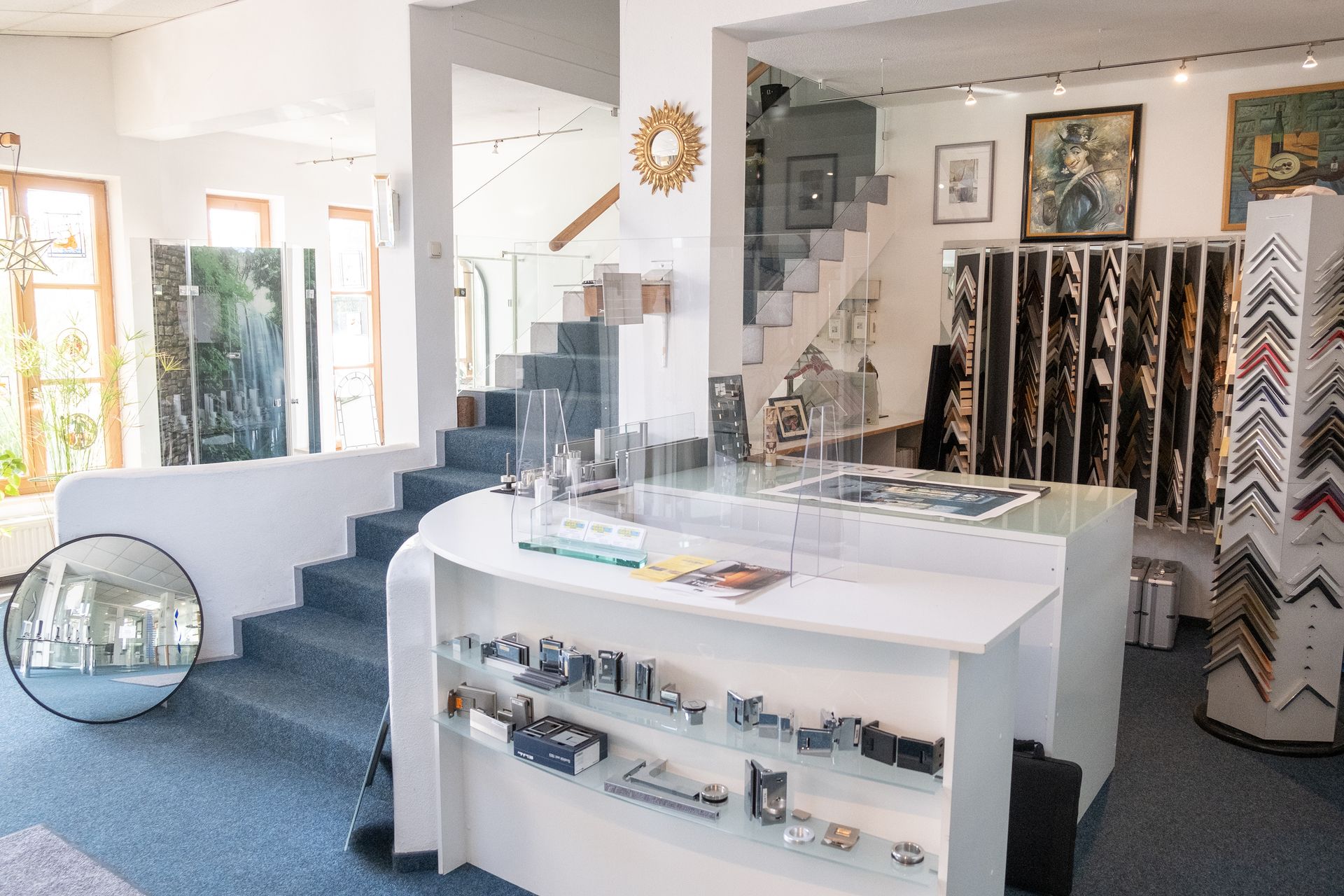 Innenraum einer Kunstgalerie mit Ausstellungstheke, Rahmen und Kunstwerken. Eine Treppe führt in die obere Etage.